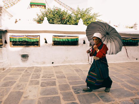 An elderly woman in traditional Tibetan dress walks with a parasol outside a temple.