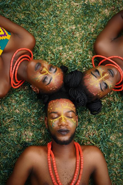 Three adults with tribal makeup and traditional beads lying on green grass, showcasing African culture.