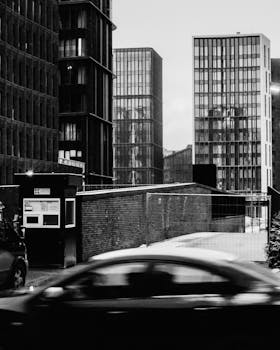 Grayscale photo of city skyscrapers and a moving car at dusk.