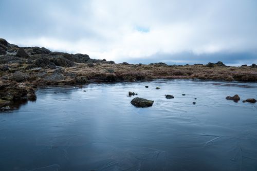 Free Peaceful winter landscape featuring a frozen lake surrounded by rocks and overcast skies. Stock Photo