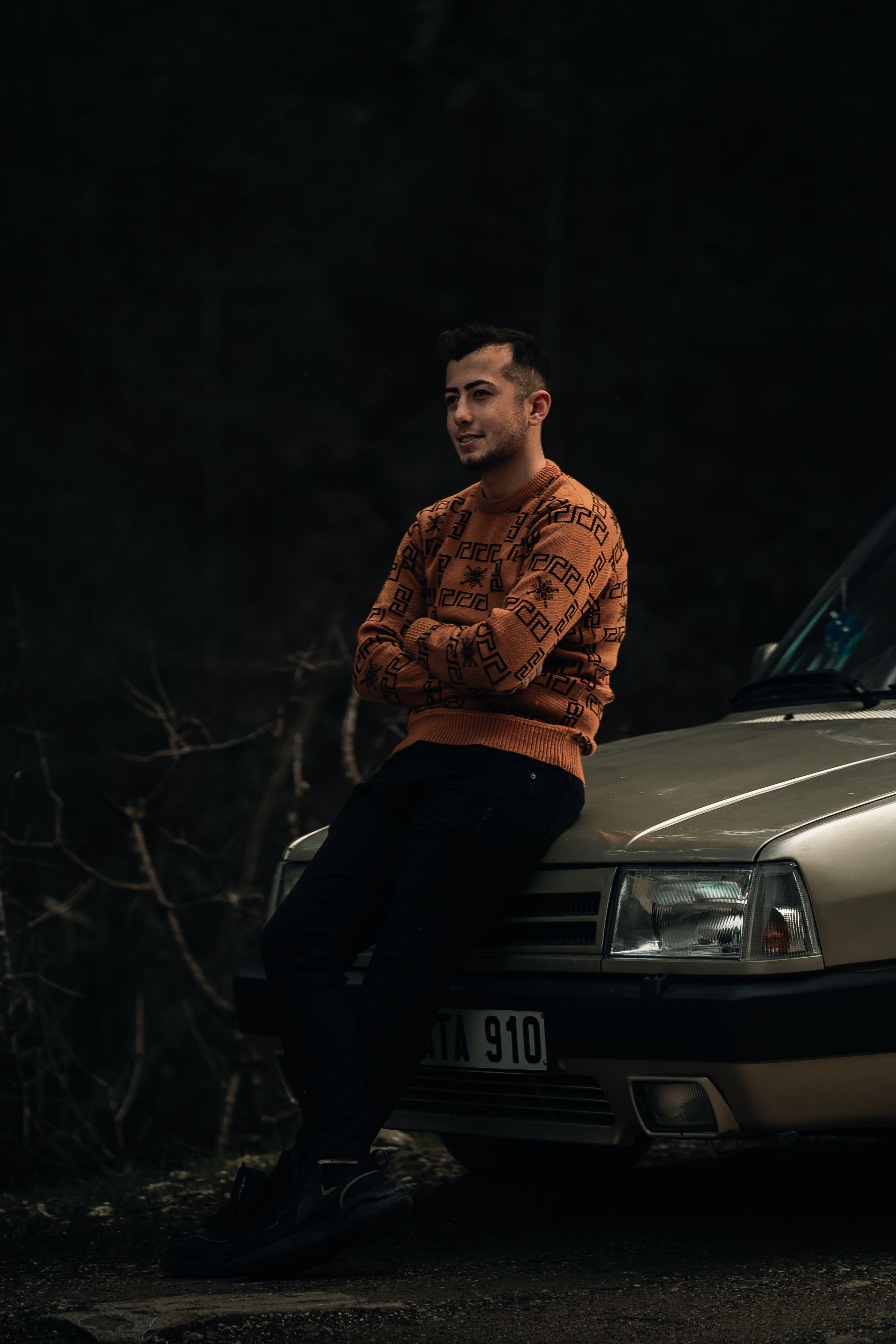 Man sits on car hood with crossed arms, wearing a brown sweater, captured in a moody and dark ambience.