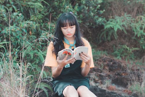 Free Asian girl enjoying a book outdoors in a natural setting, surrounded by lush greenery. Stock Photo