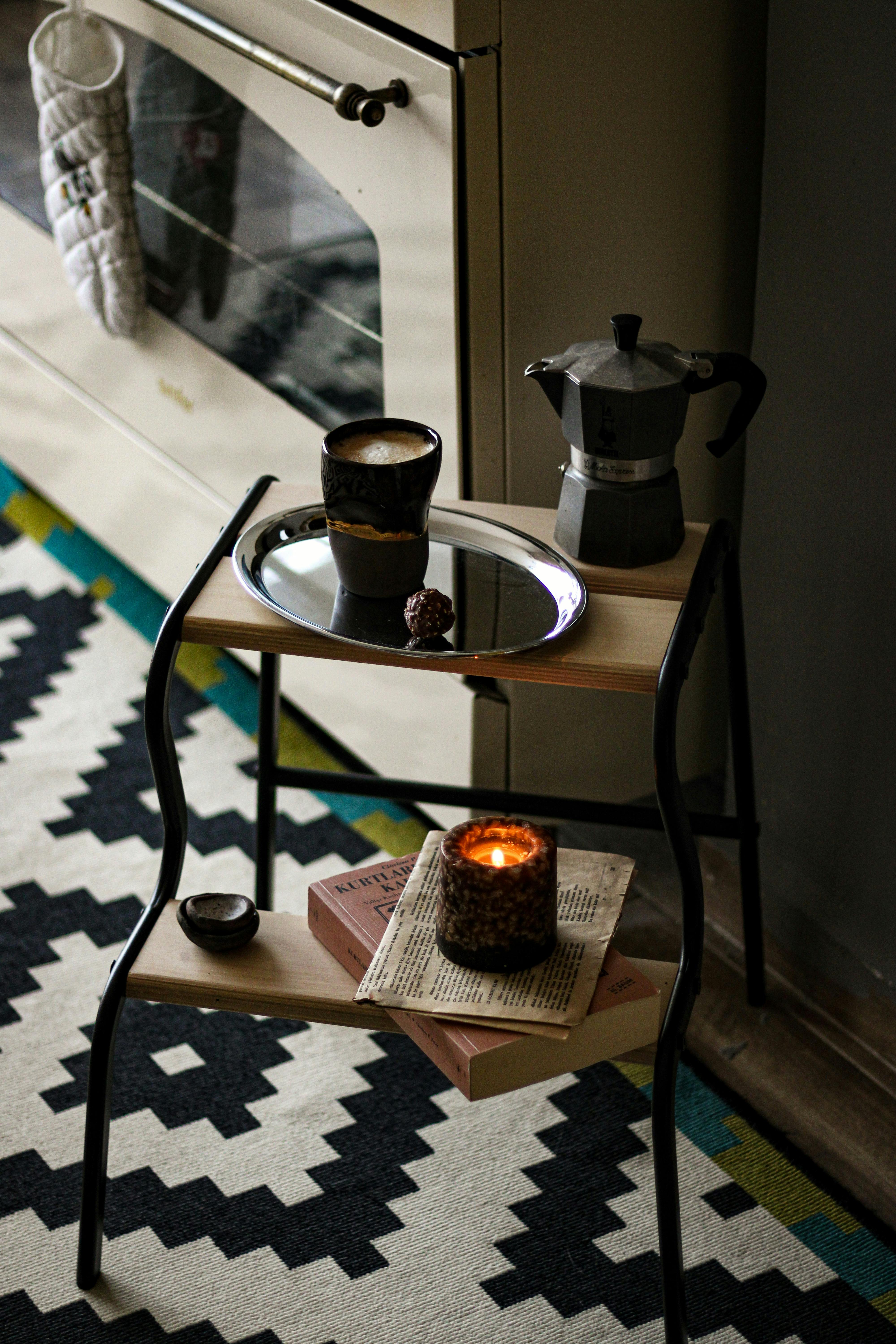 Warm indoor setting featuring a Moka pot, coffee cup, and lit candle on a small table.