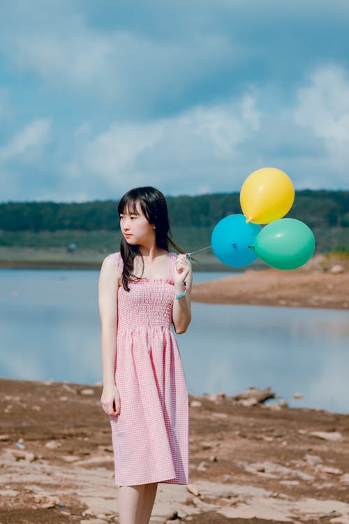 Woman Wearing Pink Dress Holding Three Balloons Near Body of Water