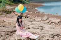 在明亮的天空下，一位年轻女子手拿五颜六色的气球，在湖边的岩石地形上享受着宁静的时刻