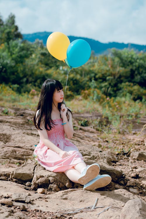 Женщина в розовом платье сидит на земле и держит два воздушных шара