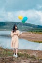 一位年轻女子身着长裙，手持五彩气球，站在风景如画的湖畔，洋溢着幸福的气息