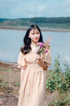 在一个阳光明媚的日子里，一个年轻的女人穿着格子布衣服，手拿鲜花，站在宁静的湖边