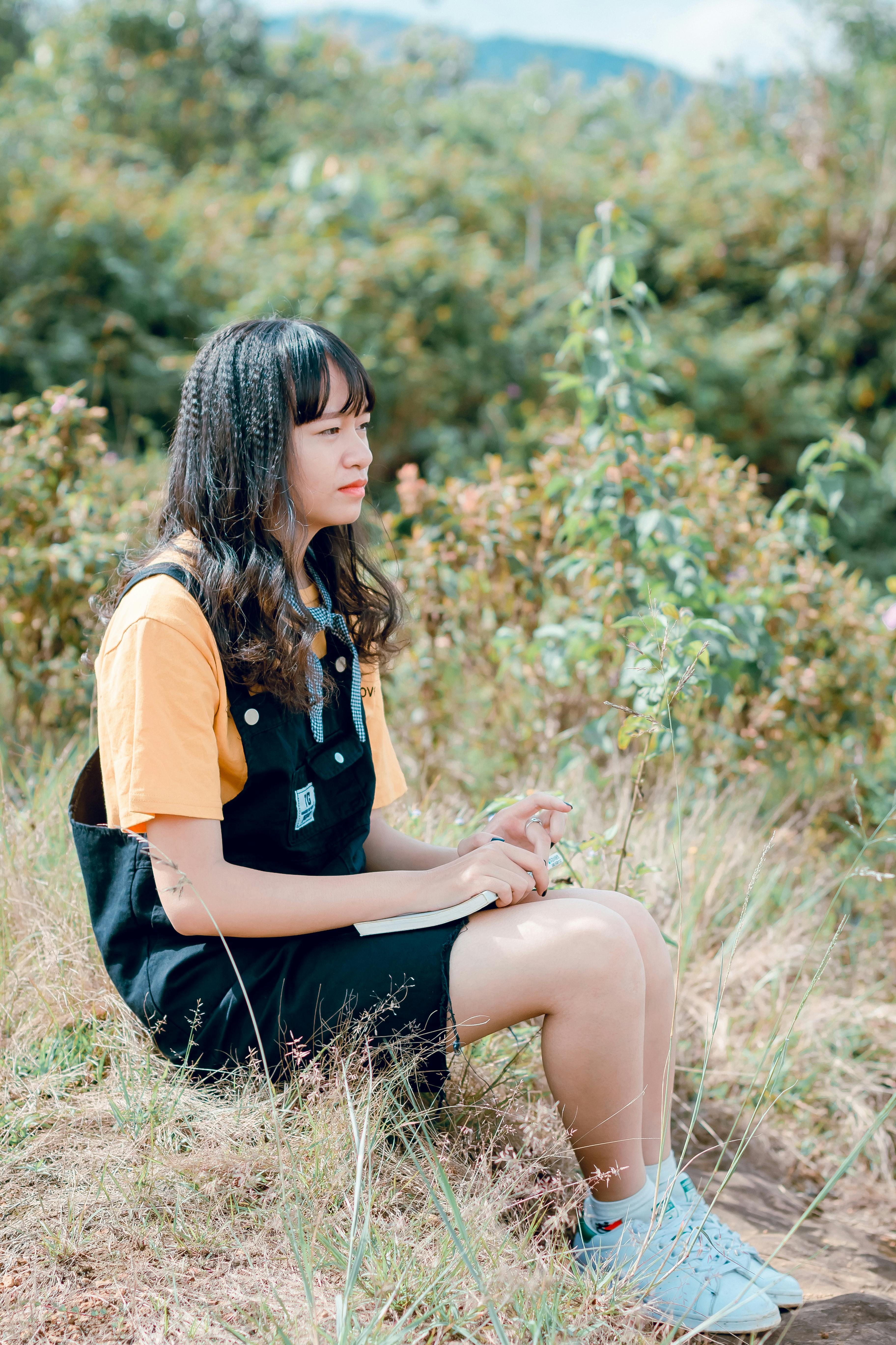 photo of woman sitting on ground surrounded by grass trees and plants