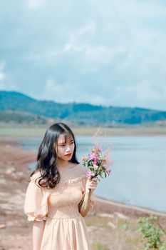 Portrait of a young woman holding flowers by a serene lake on a sunny day.