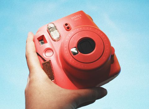 A hand holding a red Instax mini 9 camera, set against a clear blue sky.