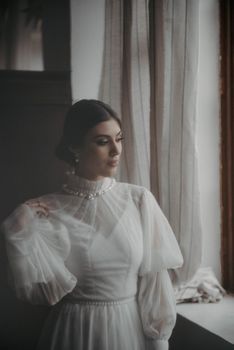 A serene portrait of a woman in a sheer white dress gazing out the window.