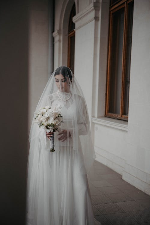 Free Beautiful Bride in White Wedding Dress and Veil Looking at Bouquet of Flowers  Stock Photo