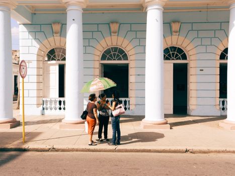 A group of women with an umbrella gather outside a colonial-style building.