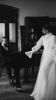 Elegant woman in white gown standing by a pianist in a vintage setting.