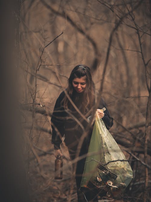 Woman Holding a Brown Bottle and a Plastic Bag in the Woods