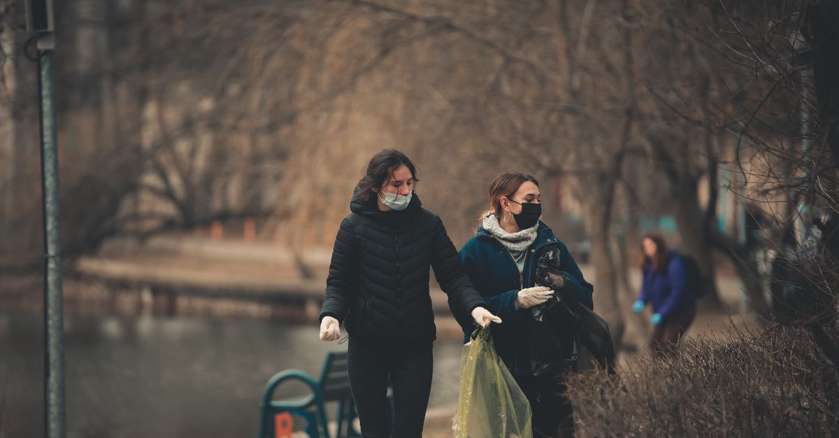 Women volunteering in autumn park cleanup, wearing face masks and gloves, holding garbage bags.