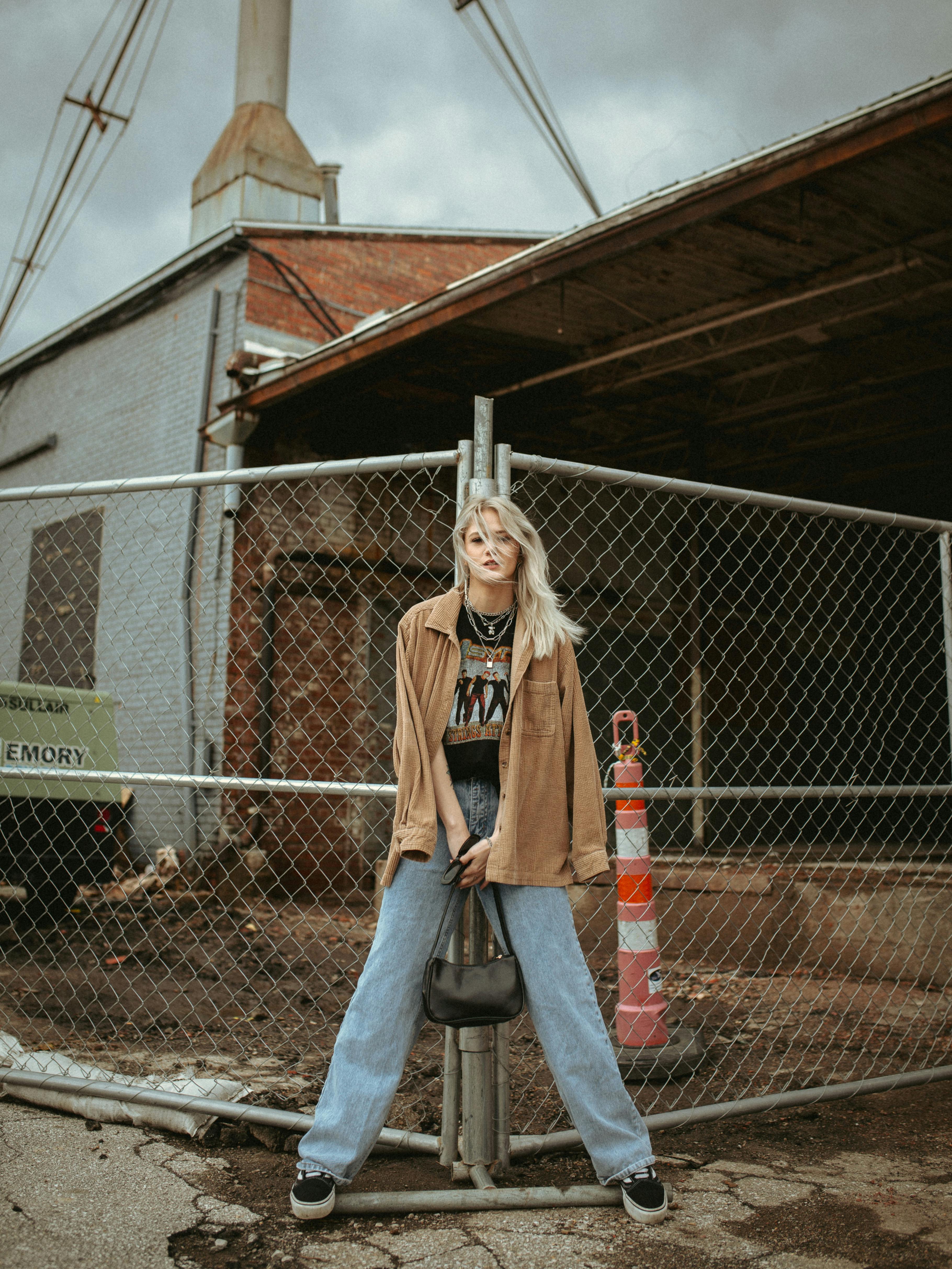 young blond woman in jeans leaning against grid metal fencing