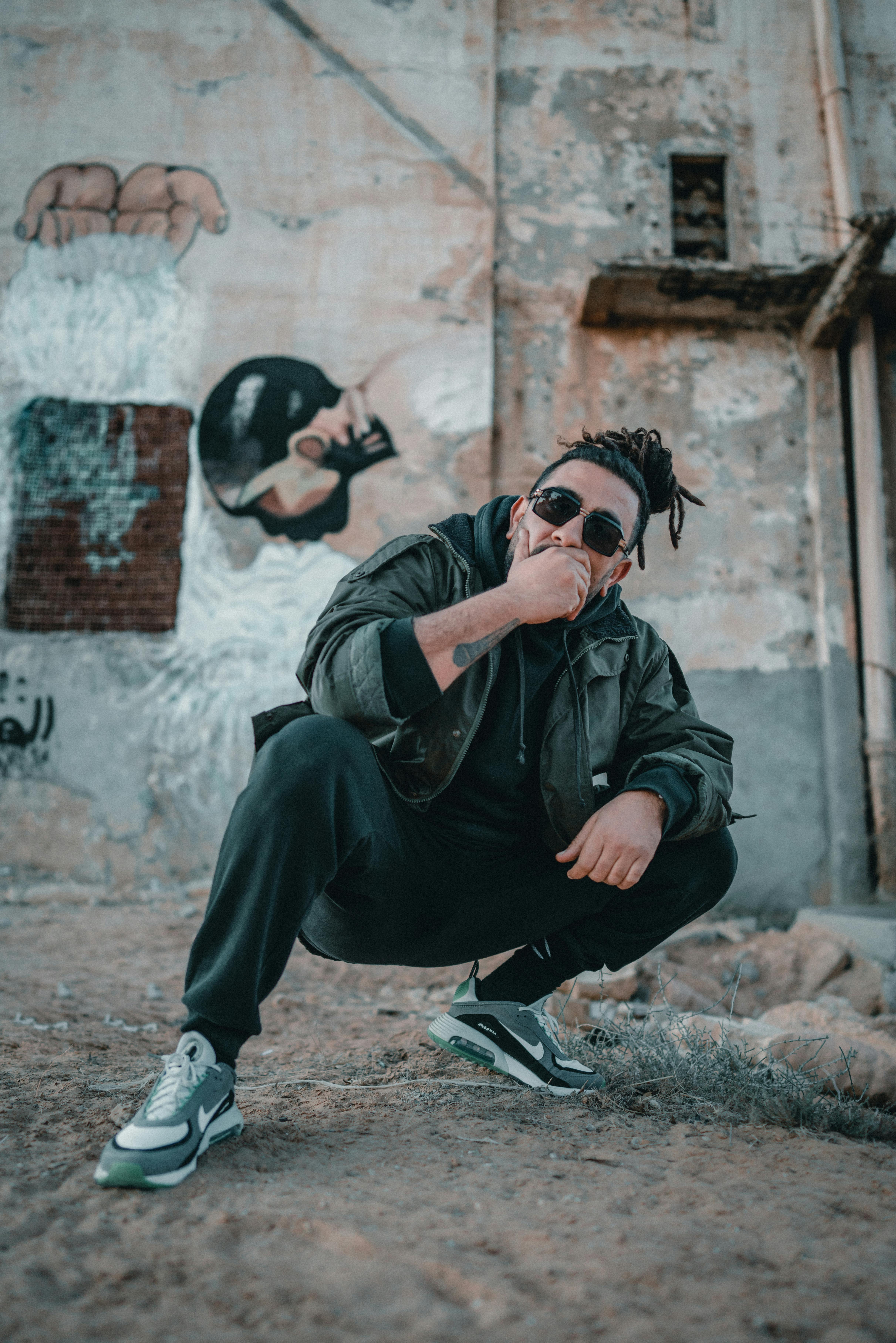 adult man in black jacket and sport pants crouching in slum backyard