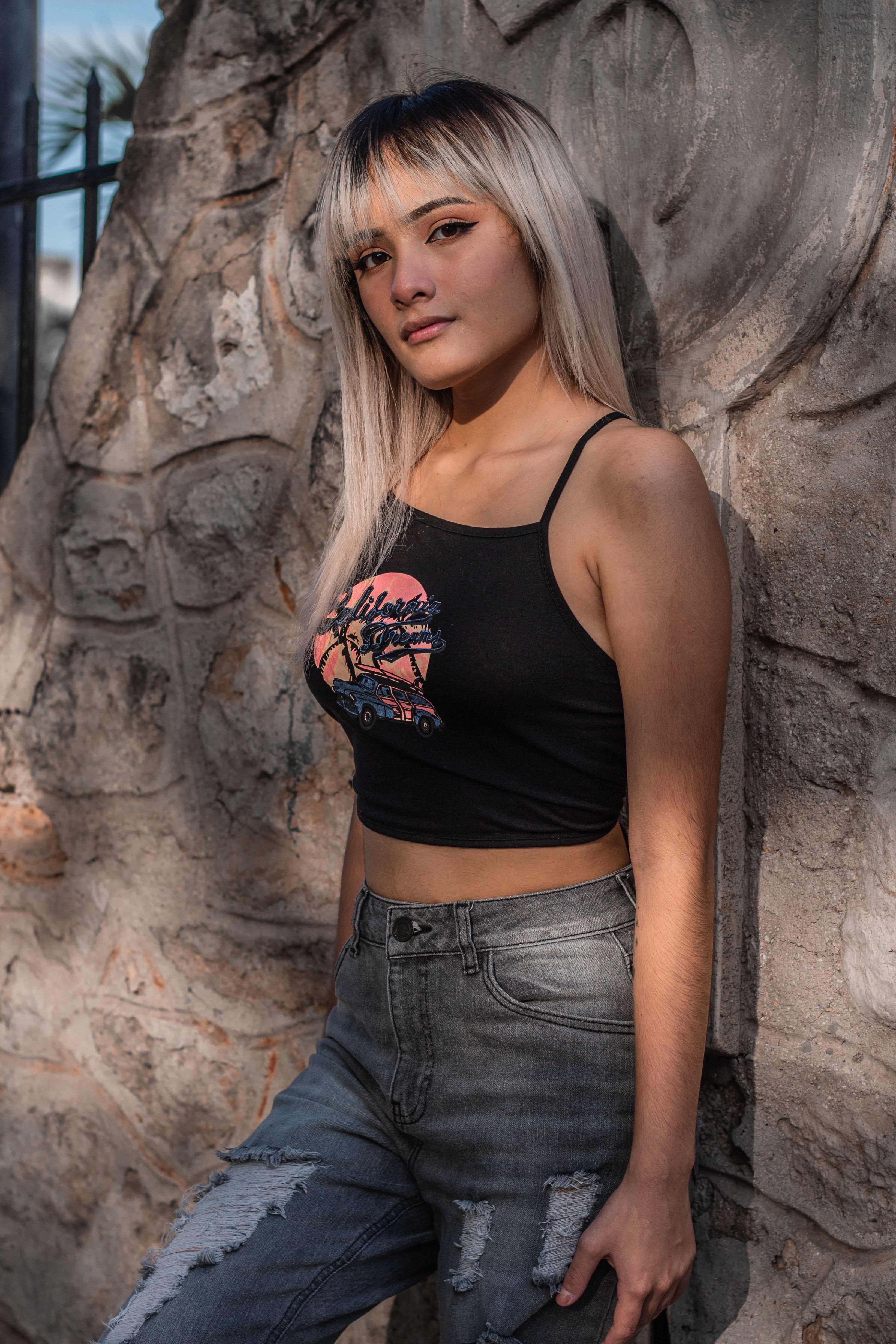 Stylish young woman in a crop top poses against a textured stone wall, illuminated by natural sunlight.
