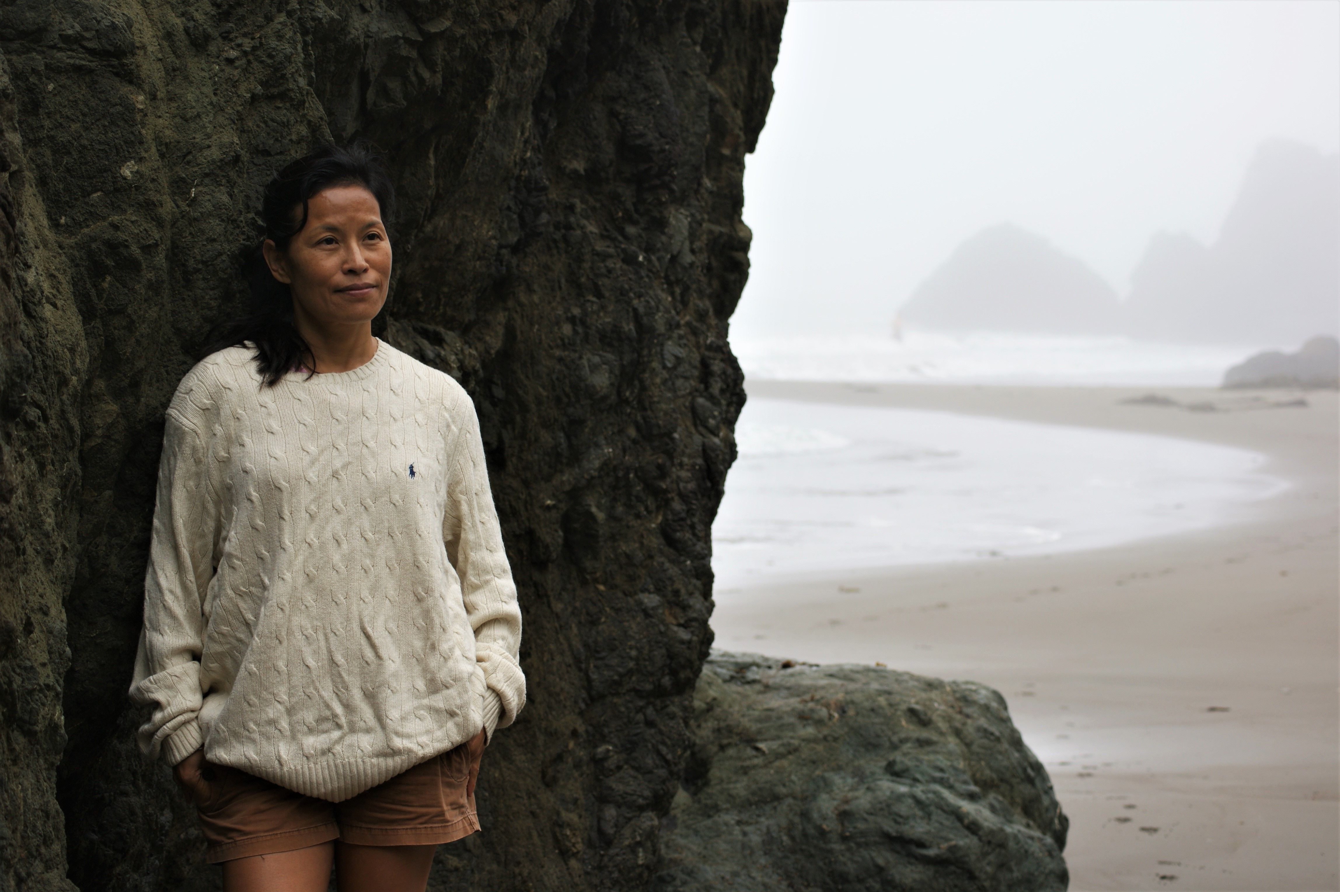 Asian woman in a cozy sweater stands by a foggy beach in Northern California, creating a serene mood.