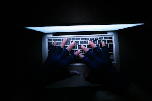 Top view of hands typing on a laptop keyboard in dim light, with gloves on.