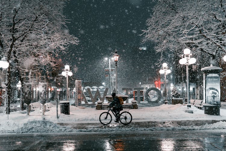 A Person Riding A Bike On The Street At Night While Snowing