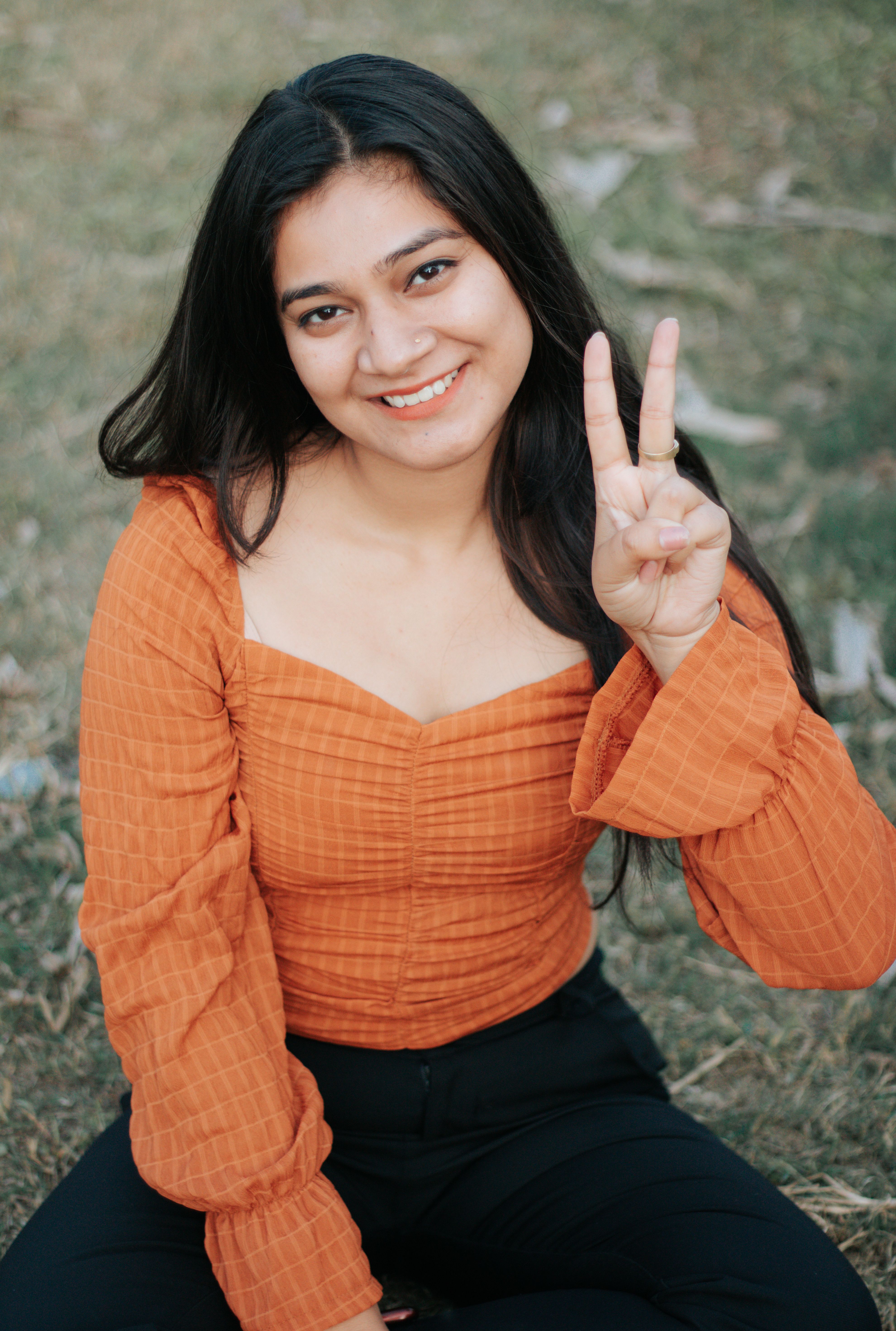 Cheerful woman with long hair in an orange top making a peace sign gesture while sitting outdoors.