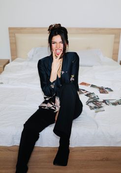 A cheerful woman in a blazer playfully sitting on a bed surrounded by photos.