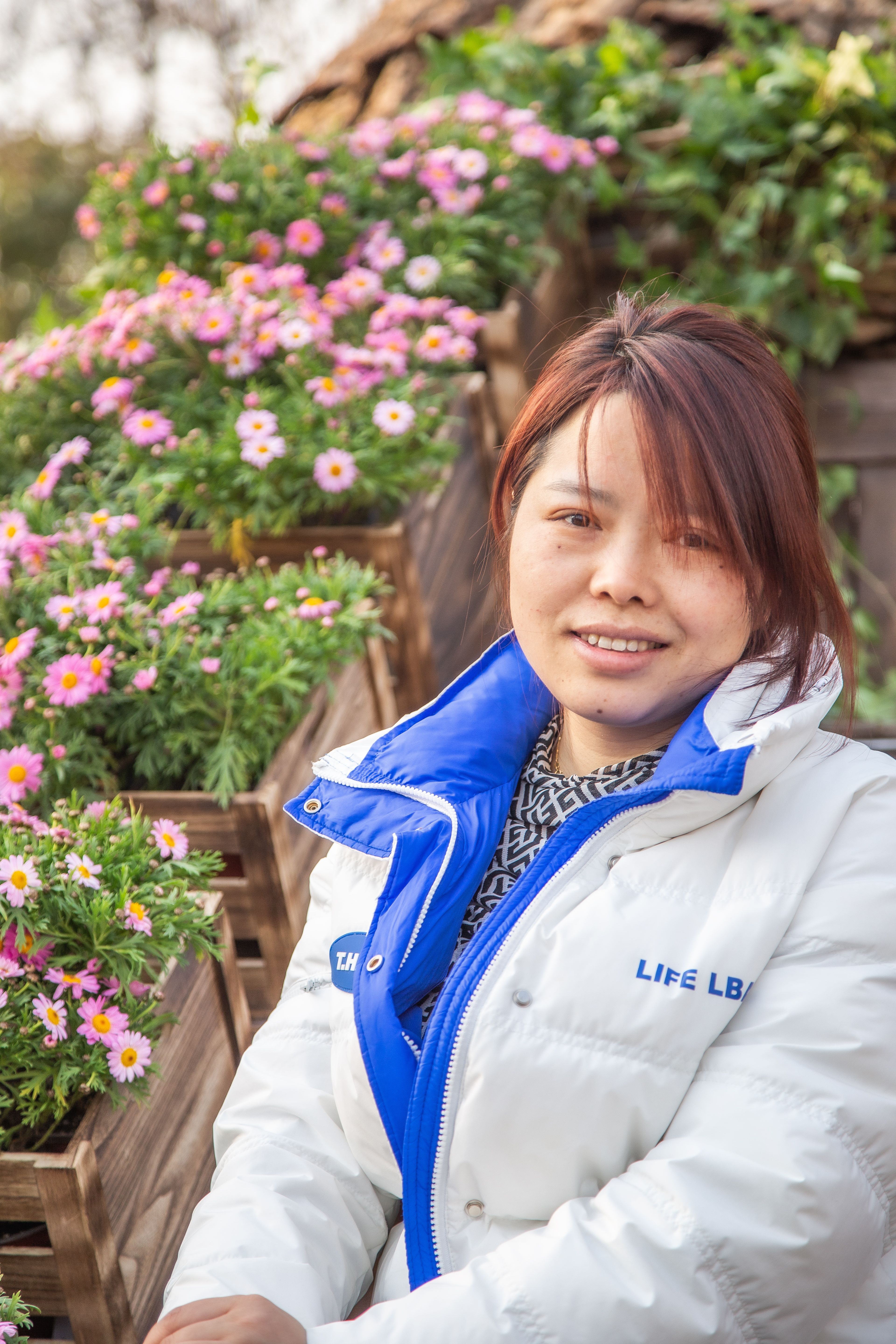 An Asian woman in a white jacket smiles outdoors with blooming flowers.