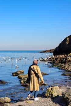 A woman wearing a hijab strolls by a scenic seashore with a flock of seagulls under a bright blue sky.