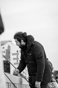 Man in a jacket fishing from a railing in monochrome setting.