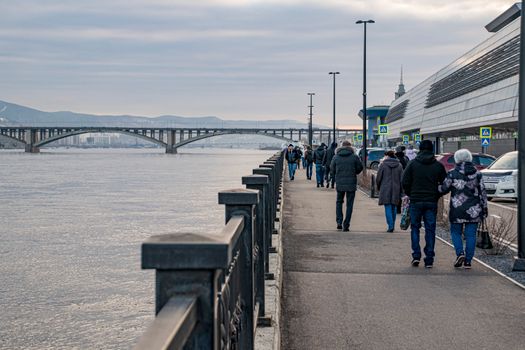 People strolling by a river under a cloudy sky, metal railings, and a bridge in the distance.