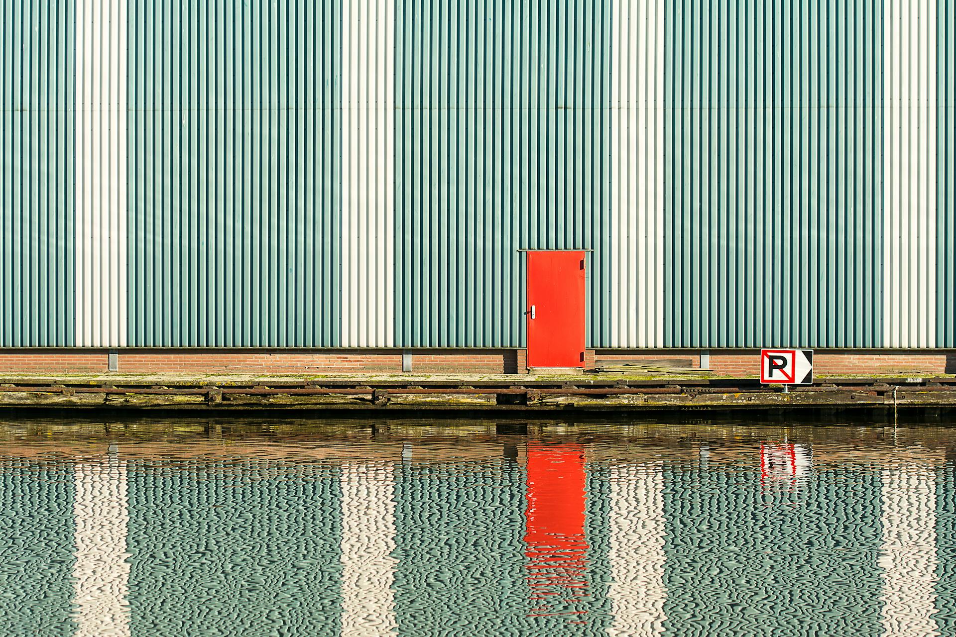 A red door on an industrial striped wall with water reflection and no parking sign.