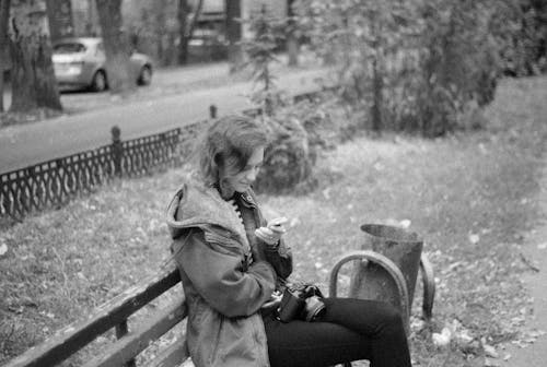 Free Grayscale Photo of a Woman Looking at Her Cellphone While Sitting on the Bench Stock Photo