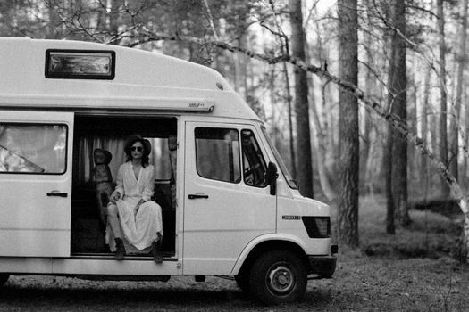 A woman sitting in a parked van amidst trees, captured in black and white.