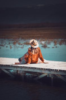 Woman in hat lounging on a pier overlooking a tranquil lake during the day.