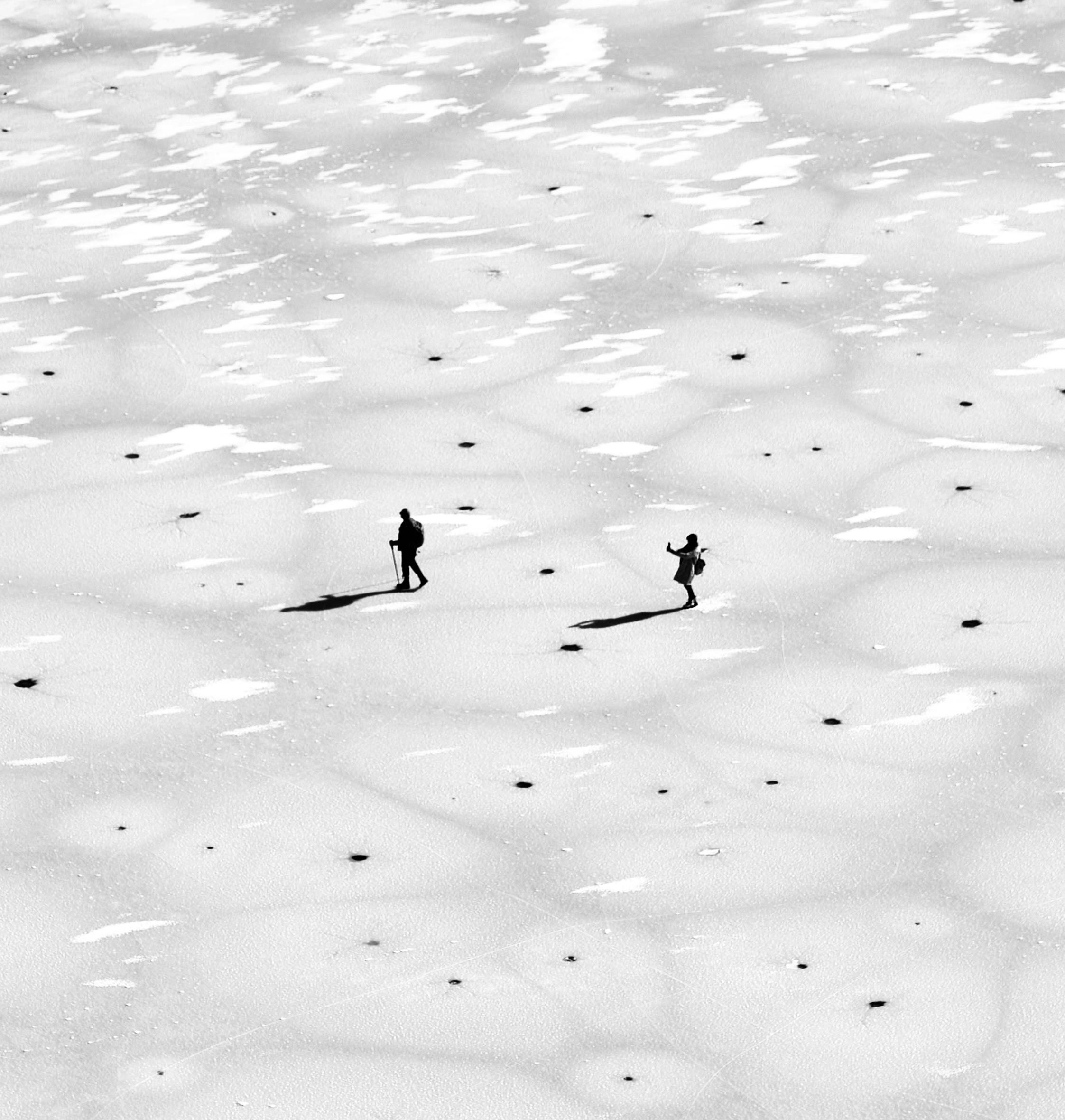 Two people traverse a snowy field in Shëngjergj, Albania, casting long shadows in black and white.