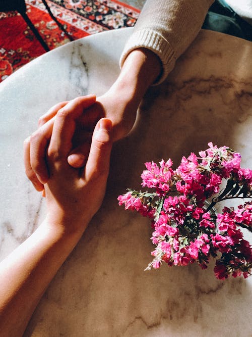 People Holding Each Others Hands while Resting on a Table Beside a Vase with Pink Flowers