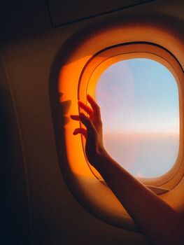 Hand reaches towards a sunset view from an airplane window, capturing a travel moment.