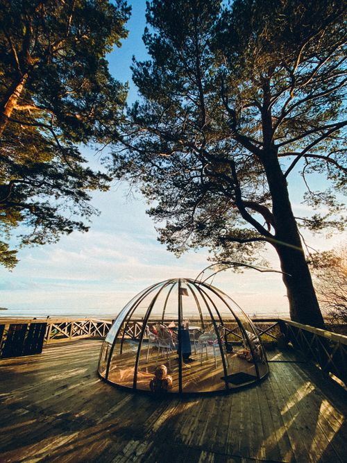 Free A picturesque glass dome sitting on a wooden terrace surrounded by tall trees and overlooking a peaceful lake at sunset. Stock Photo