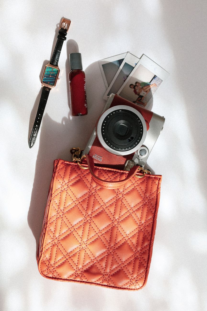 Flat lay of a polaroid camera, orange designer bag, wristwatch, and perfume on a white background.. flat, polaroid, camera