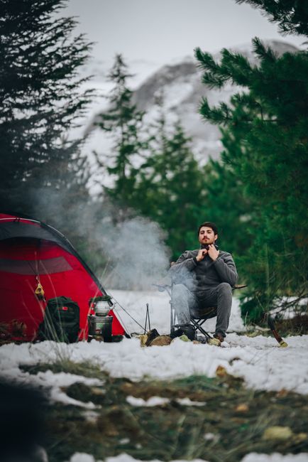 Snowy campsite with a tent, backpack, and warm glow from a lantern among pine trees