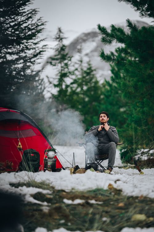Free A man sitting by a red tent during a winter camping trip in a snowy forest. Stock Photo