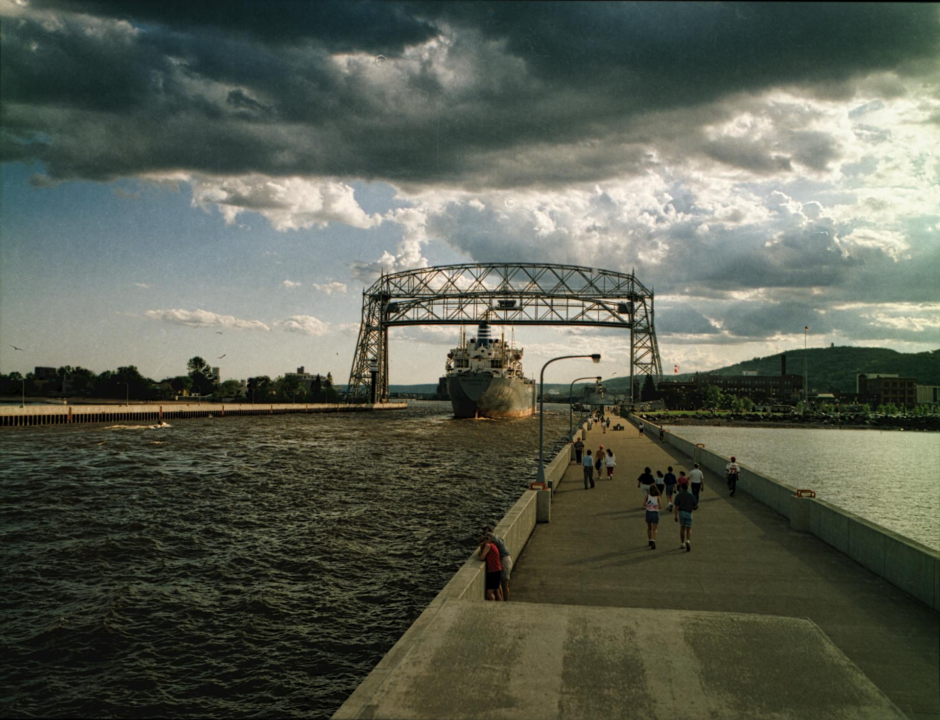 A scenic view of Duluth's Aerial Lift Bridge with ship passing and people strolling on a cloudy day.