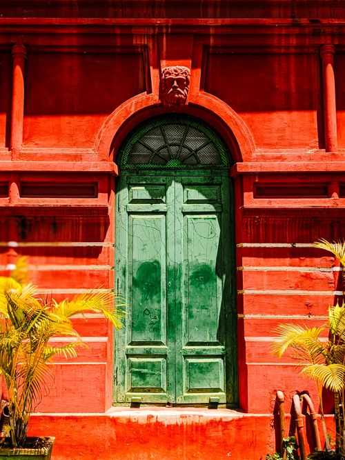 Free A vibrant green door set against a striking red wall with tropical plants in India. Stock Photo