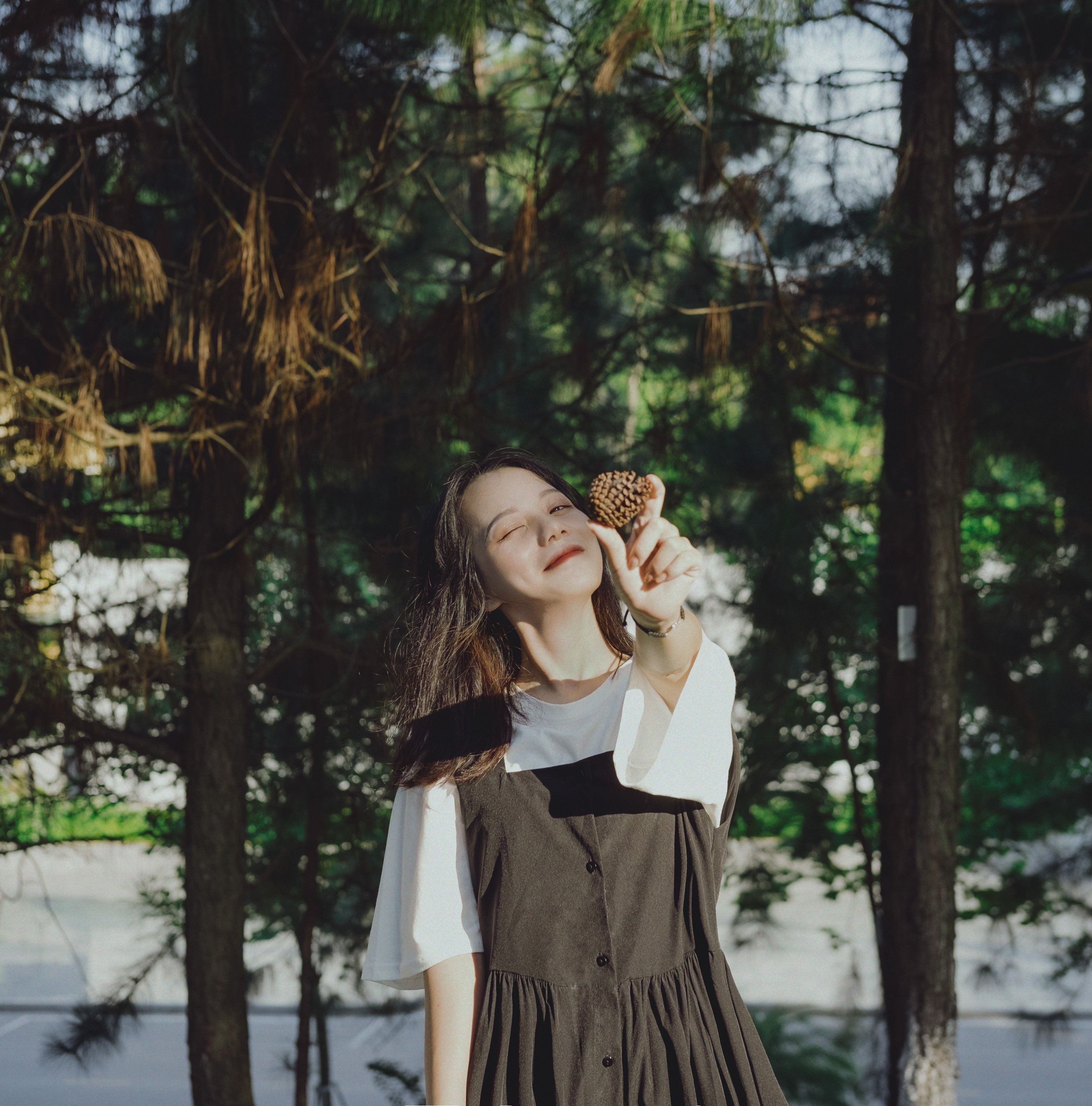 A woman joyfully holds a pine cone in a sunlit forest, embracing nature.