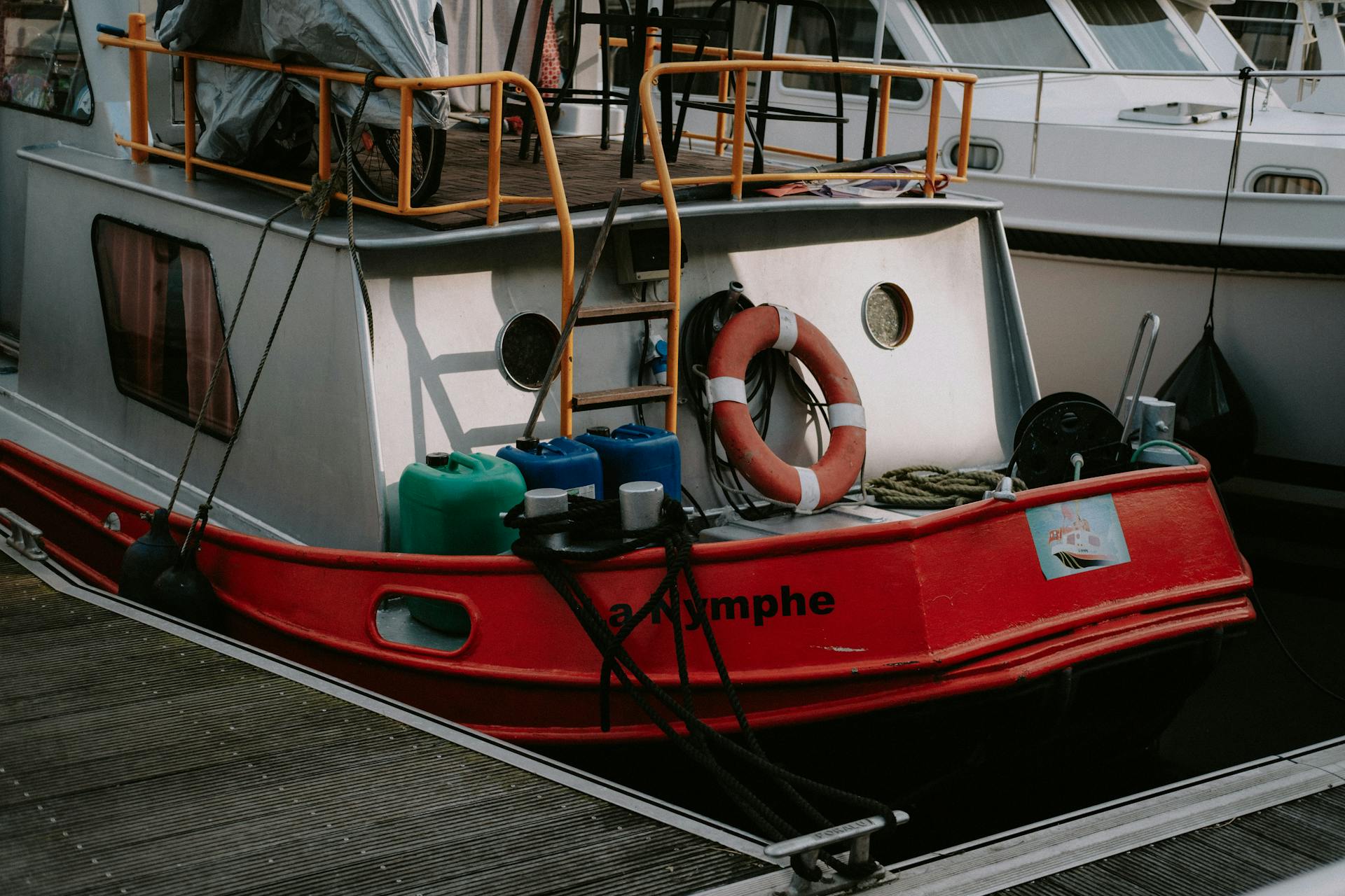 Colorful houseboat docked in a marina with lifebuoy and plastic containers visible on deck.