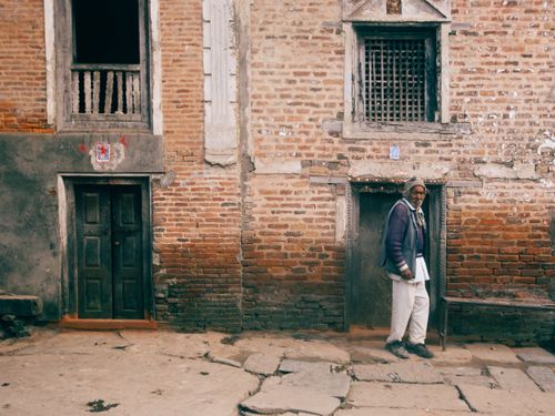 Free An elderly man stands by a weathered brick building with wooden doors, showcasing urban decay and heritage. Stock Photo