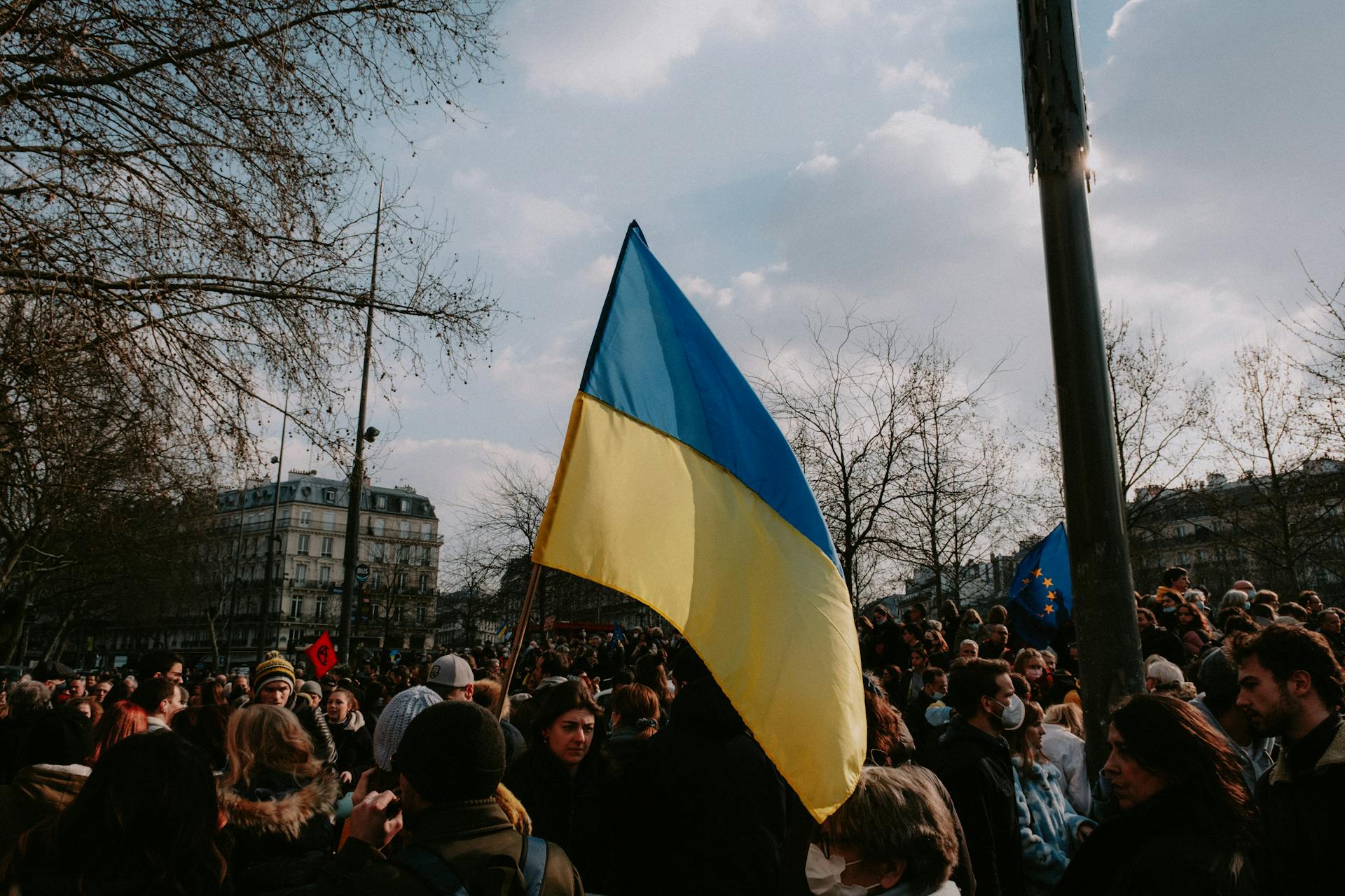 Crowd gathered for a protest with Ukrainian and EU flags under a cloudy sky.